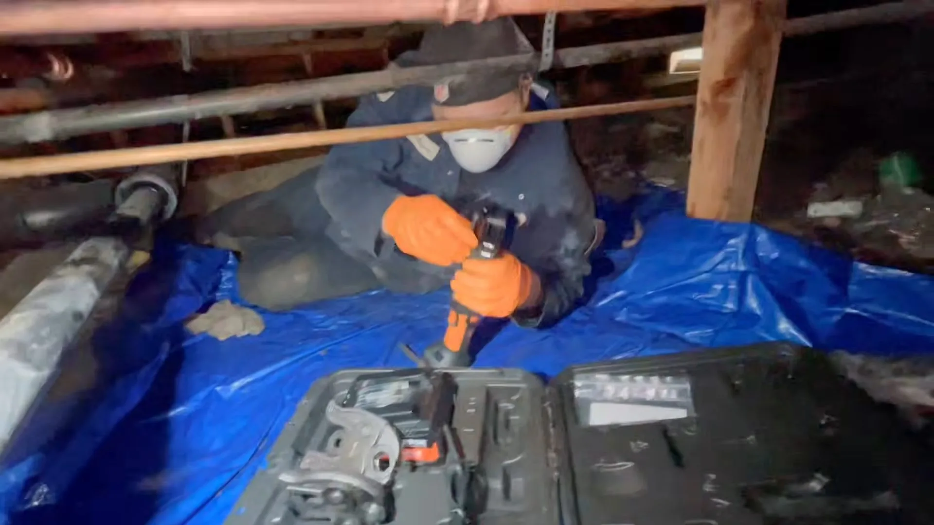 Worker in a dust mask and orange gloves kneeling on a blue tarp in a crawl space, with a toolbox open beside them.