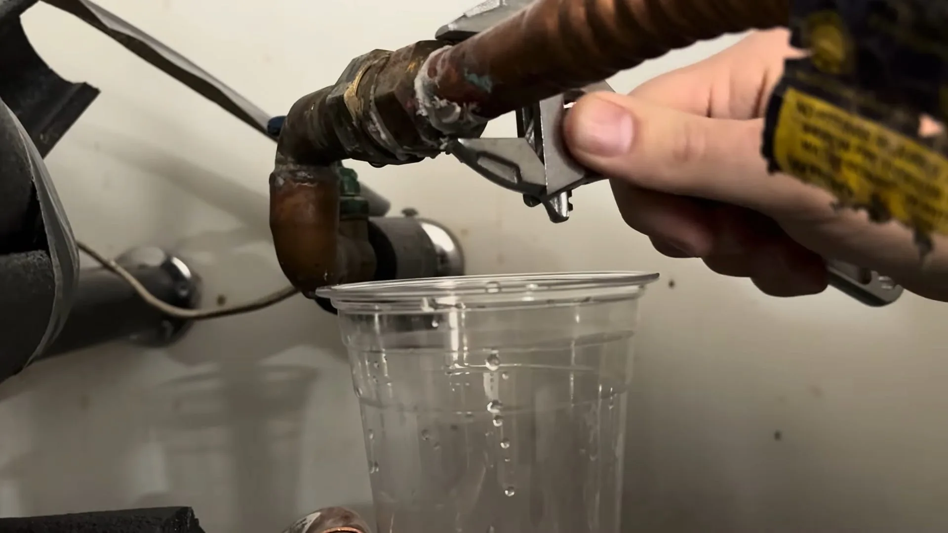 Hand holding a wrench on a corroded copper and brass fitting, with a clear cup below catching drips.